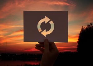 Hand holding a paper cut-out refresh icon against a sunset sky, symbolizing renewal and automation